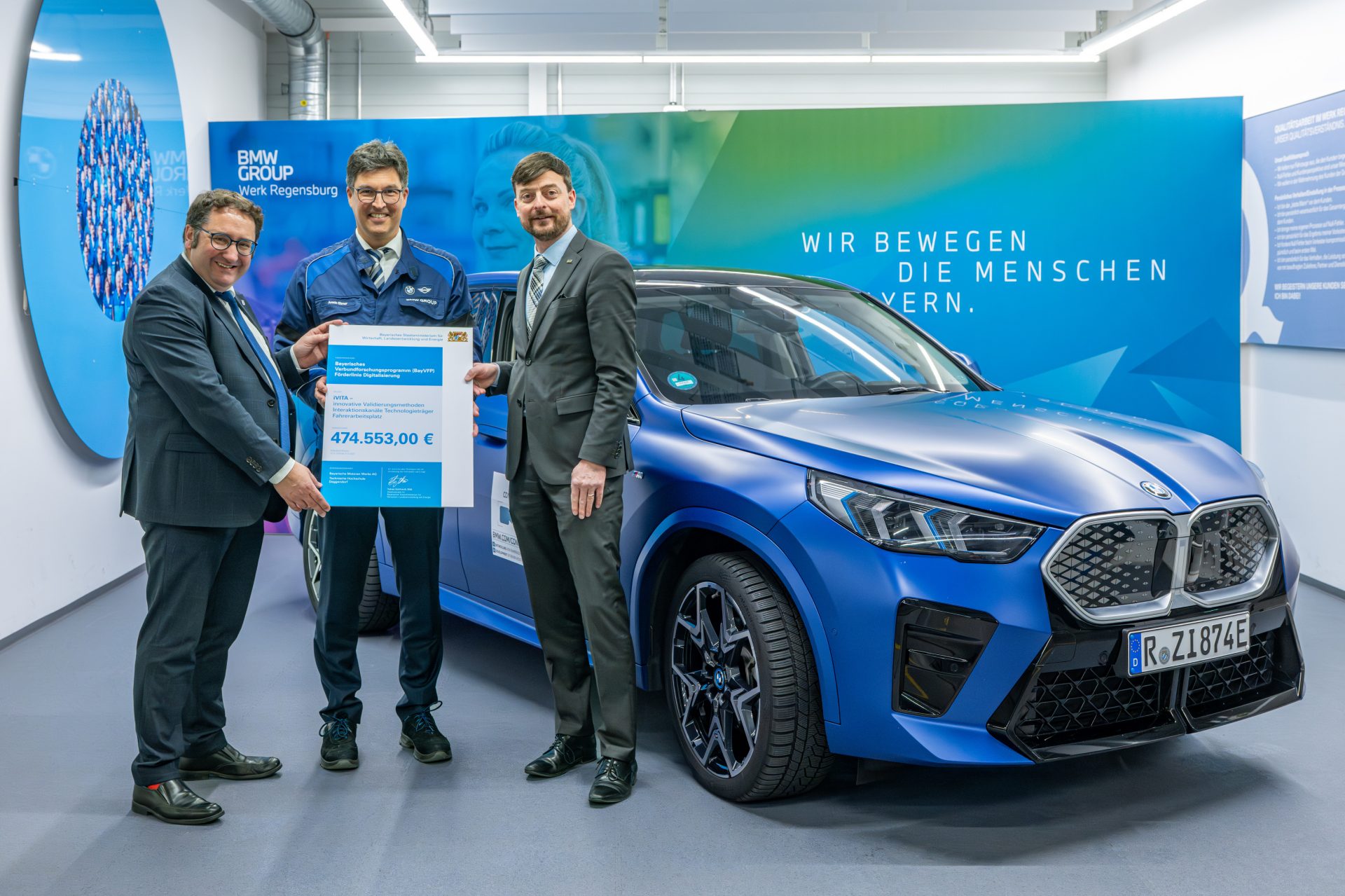 Drei Männer im Anzug stehen neben einem blauen BMW iX1 und halten einen Spendenscheck in den Händen.