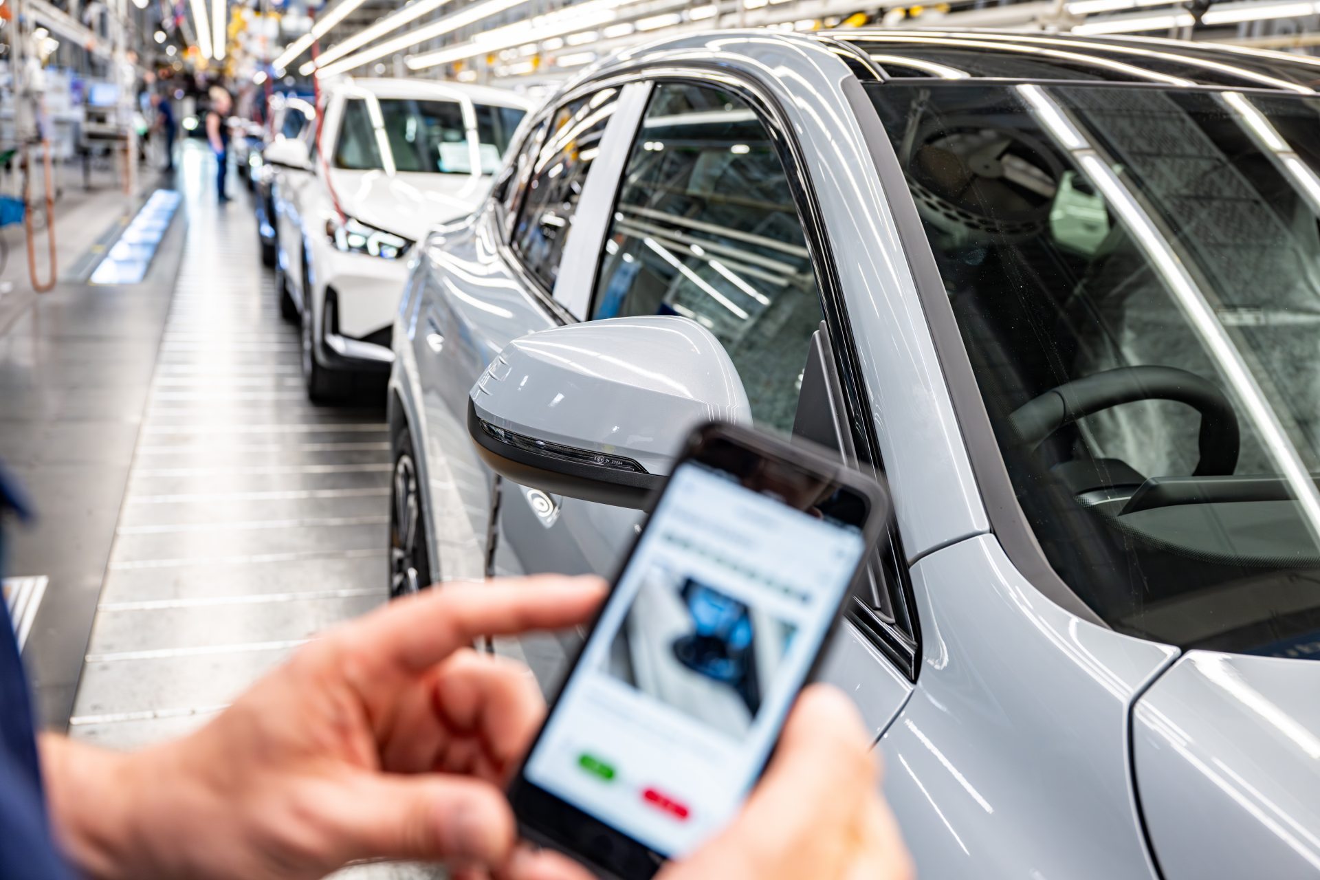 A person holds a smartphone in both hands and stands next to a vehicle on the production line.