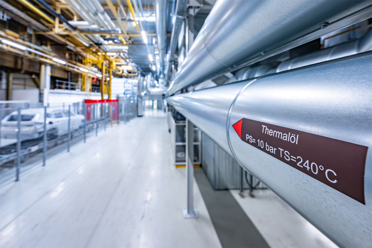 Pipeline with brown label 'Thermalöl PS= 10 bar TS=240°C' in industrial hall with metal structure and lighting.
