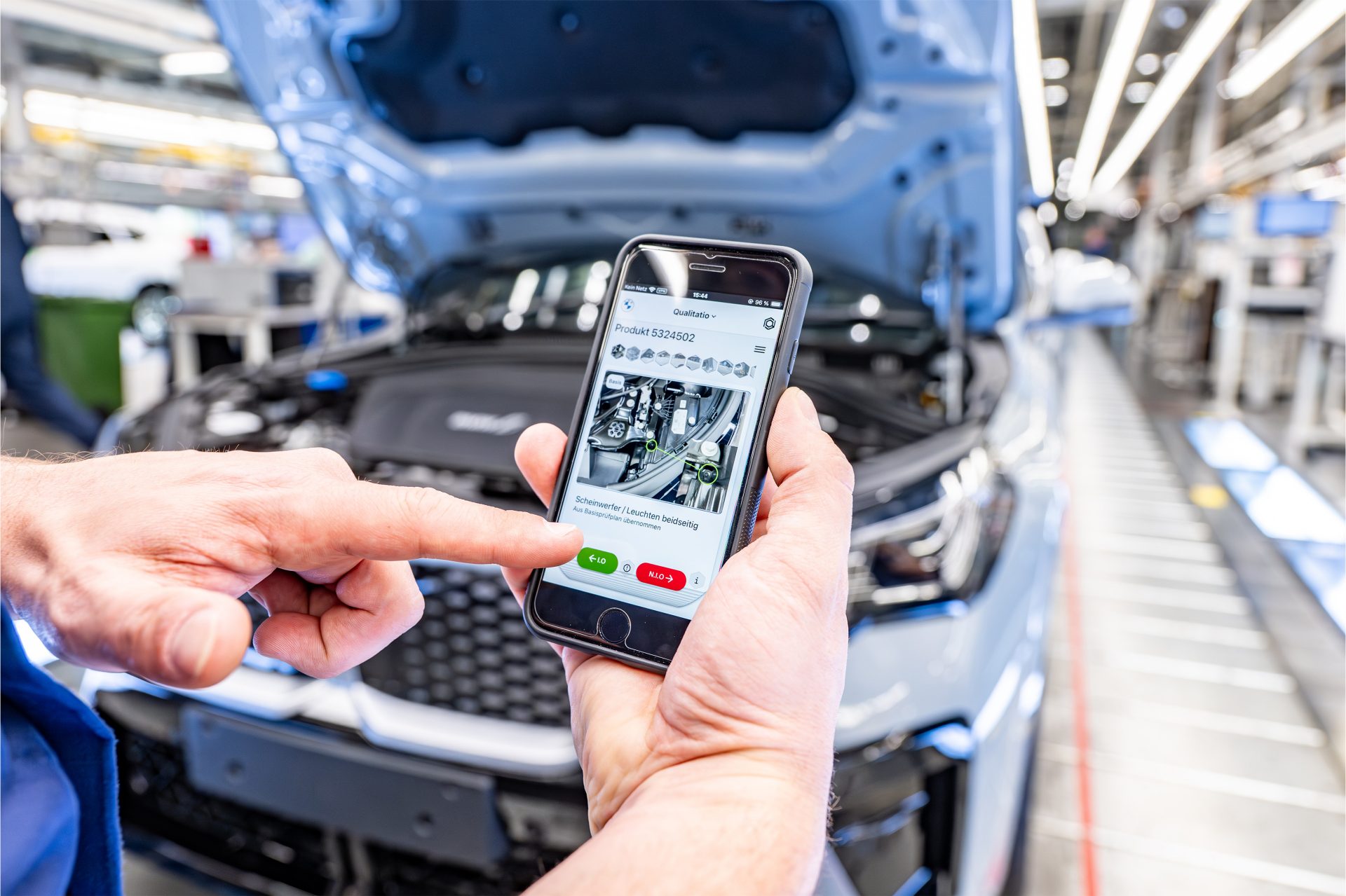 A person holds a smartphone in both hands and stands next to a vehicle on the production line.