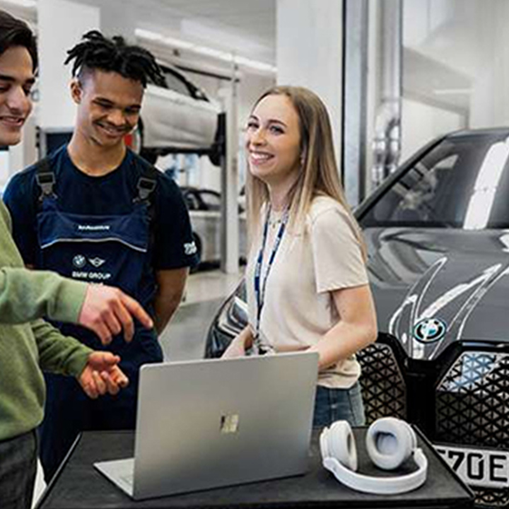 Three students stand together in a workshop in front of a BMW iX and discuss something on the computer.