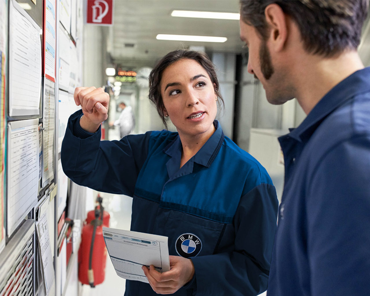 An employee at the BMW plant explains the processes to her new colleague. Both are wearing blue overalls.