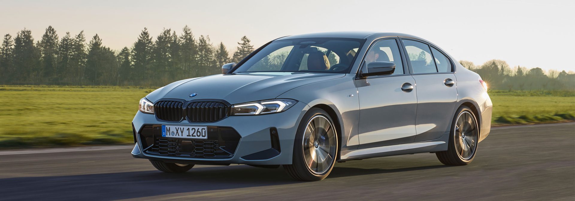 Grey BMW Series 3 driving on a road next to a wide field.