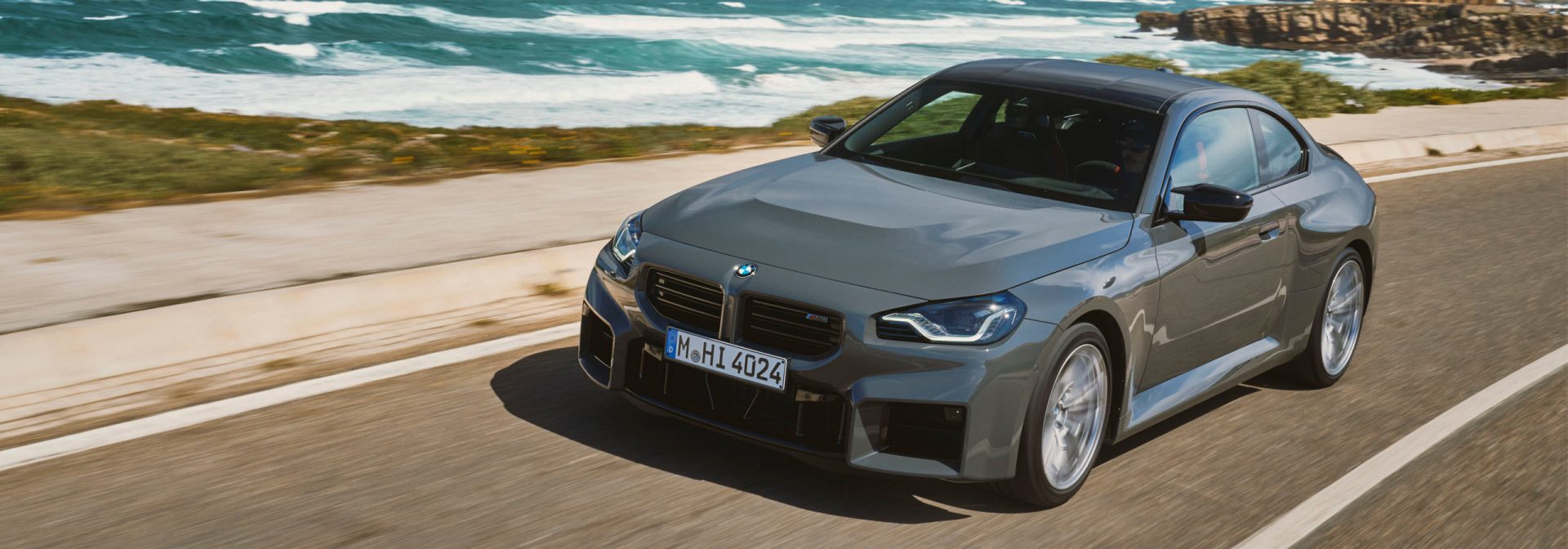 Grey BMW M2 driving on a road next to the sea.