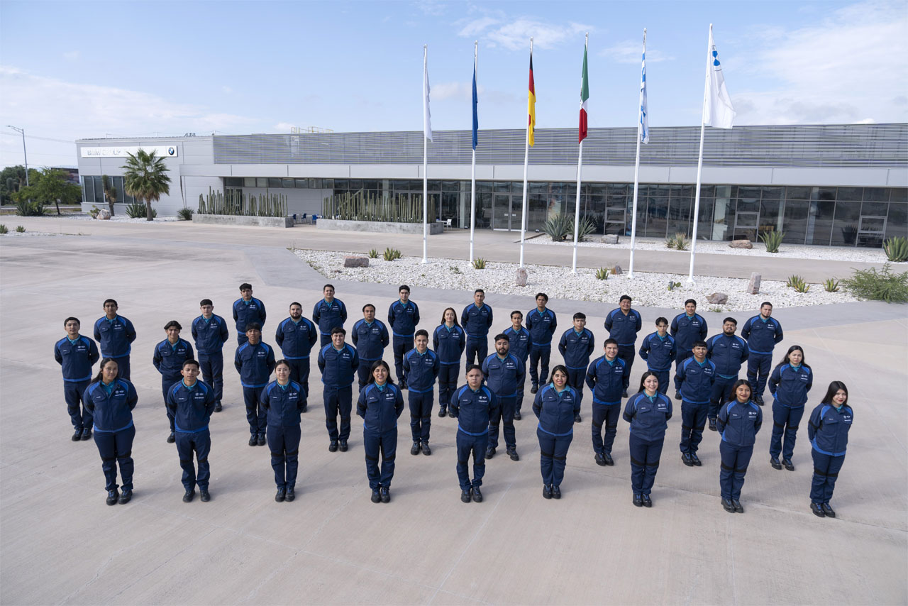 Group of 27 people in blue uniforms standing outdoors in rows on a concrete surface with five flags and a modern building in the background.