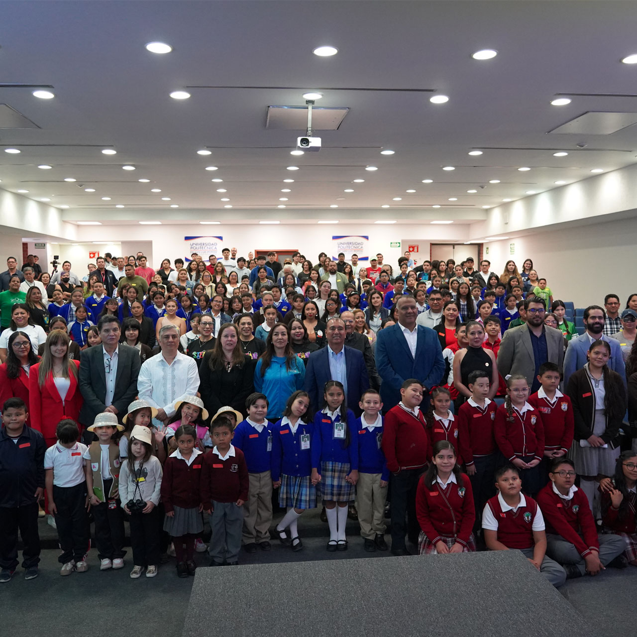 Large group of children and adults posing in a conference room with white ceiling and bright lights.