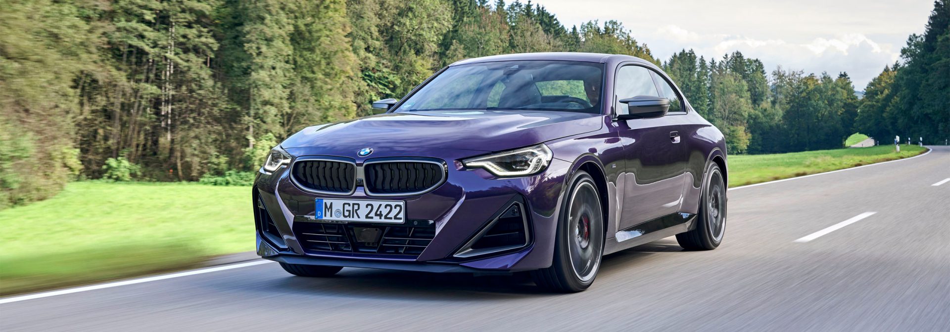 Purple BMW 2 series driving on a road through the woods.