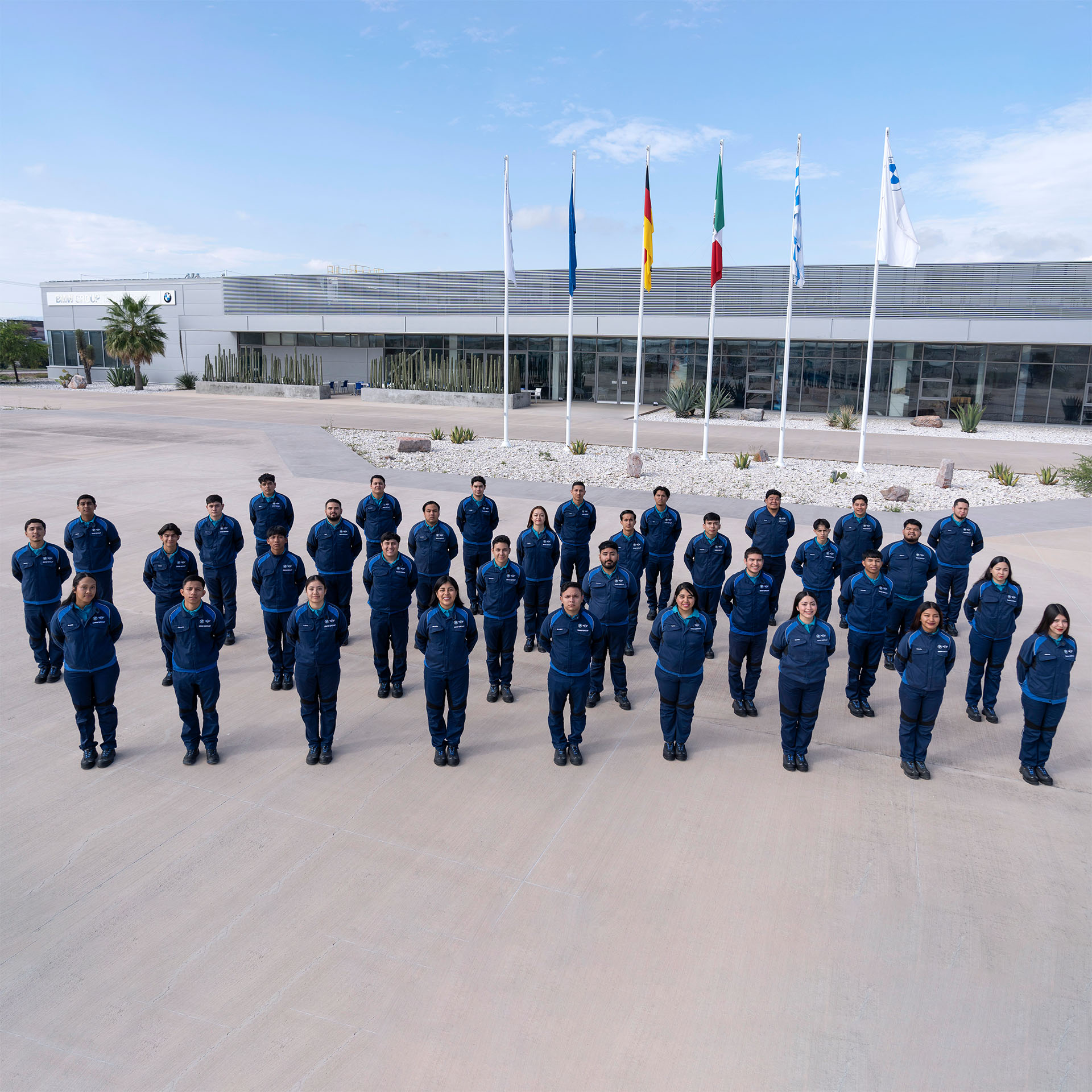Group of people in blue uniforms standing outdoors in rows on a concrete surface with five flags and a modern building in the background.