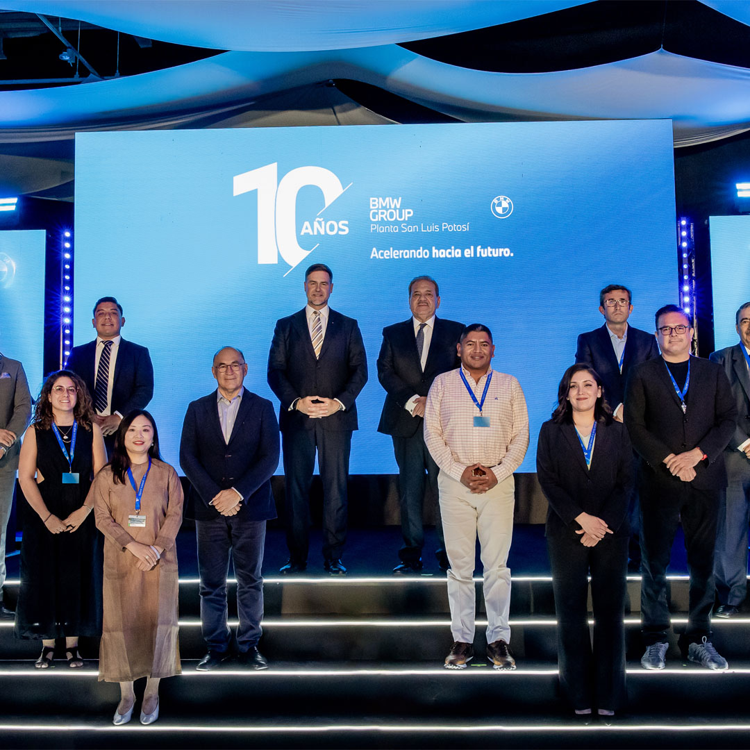 Grupo de personas con ropa formal en un escenario iluminado frente a una pantalla que muestra '10 AÑOS BMW GROUP Planta San Luis Potosí Acelerando hacia el futuro'.