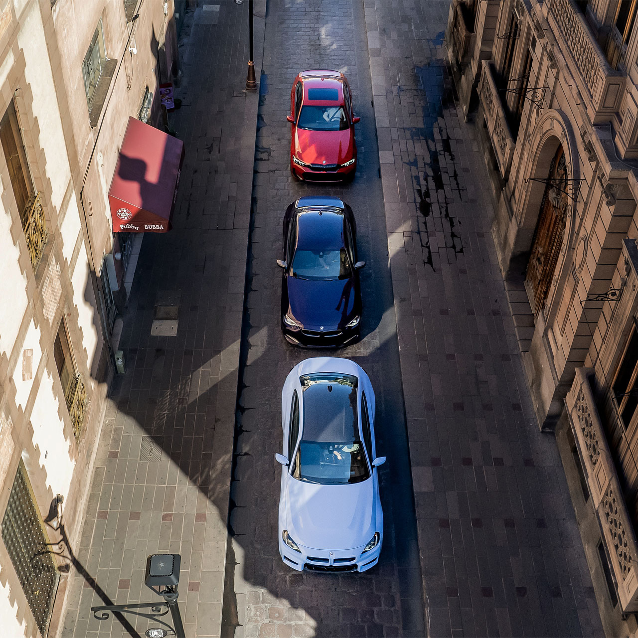 Tres coches en fila en una calle estrecha entre edificios altos vistos desde arriba.