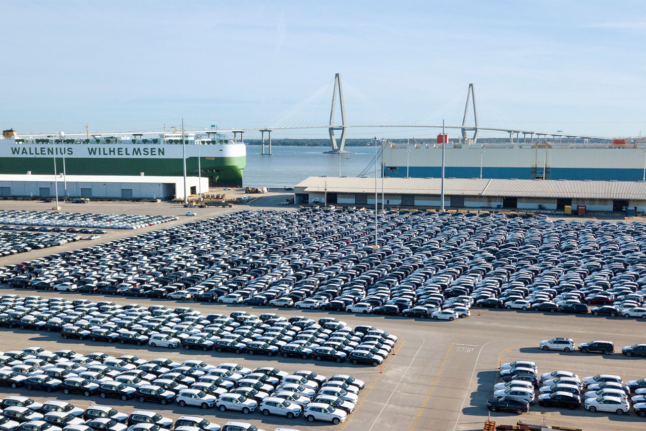 Large port area with many parked cars and a cargo ship in the background in front of a bridge