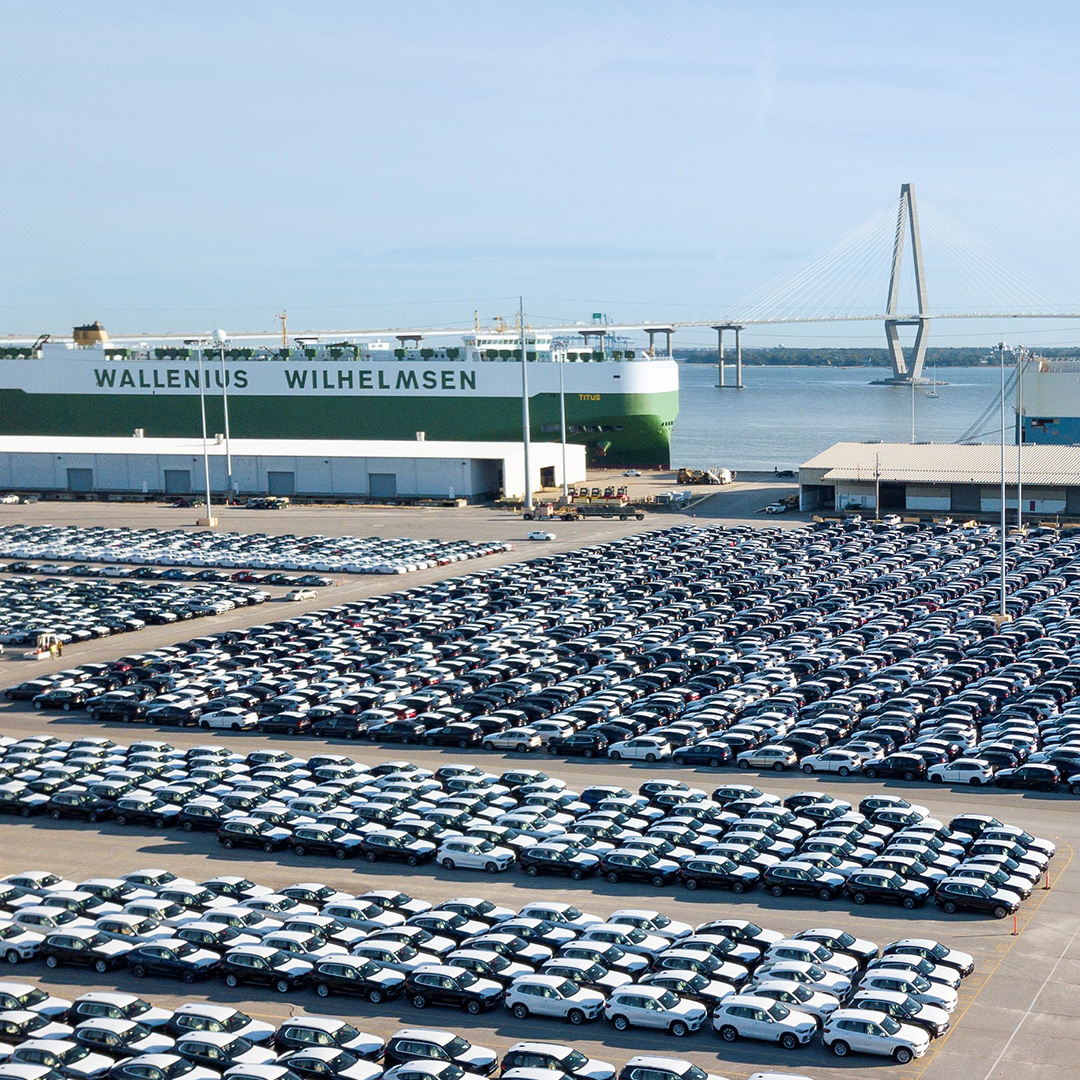 Large port area with many parked cars and a cargo ship in the background in front of a bridge