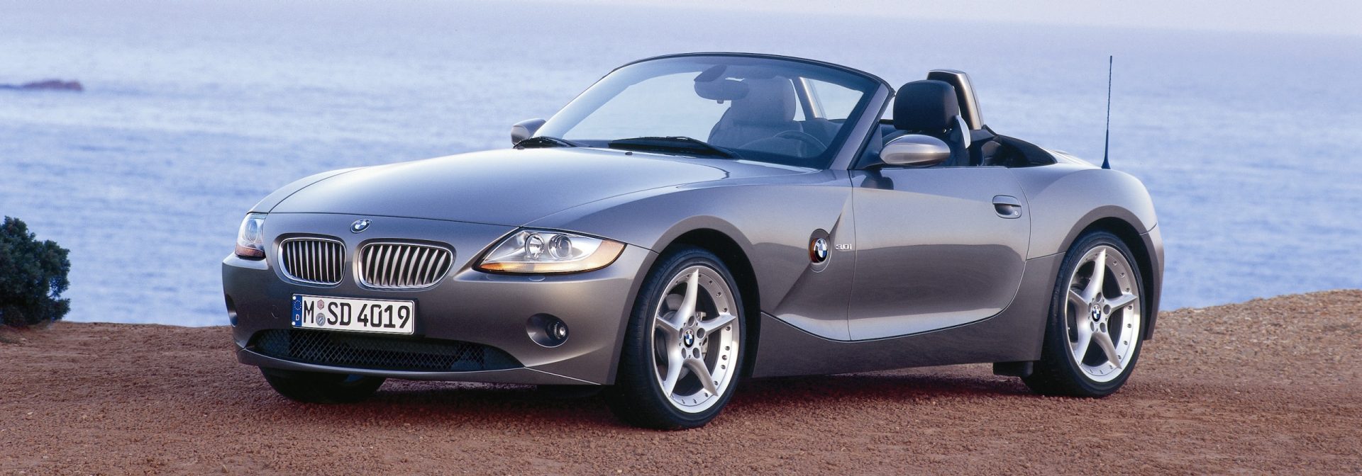 Silver BMW Z4 Roadster with open top, parked on reddish ground by the sea.