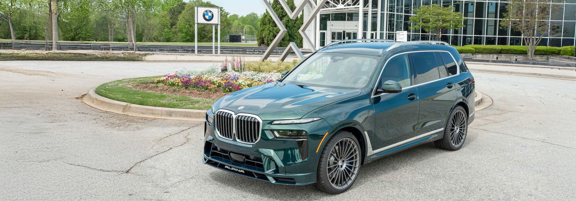 Dark green BMW Alpina XB7 SUV parked on asphalt in front of a BMW logo and a modern glass building.