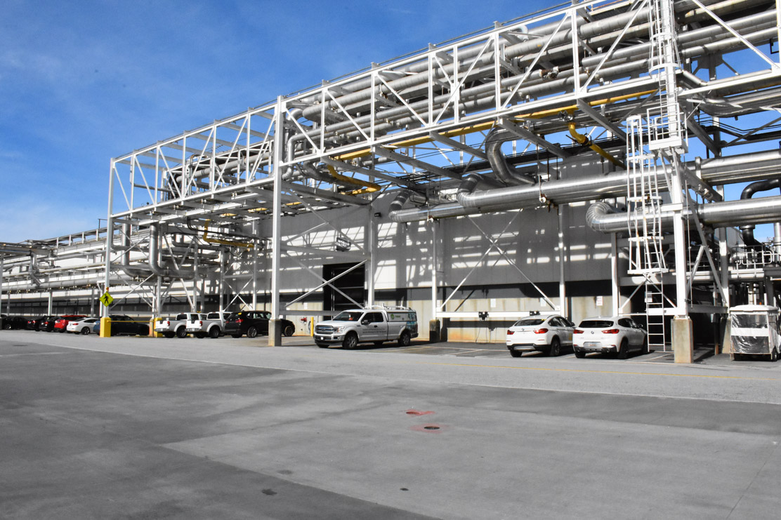 Large industrial facility with multiple white vehicles parked under a complex network of pipes and steel beams under clear sky.
