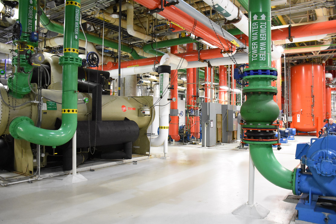 Mechanical room with large green, red, and white pipes, valves, and machinery on a light floor.