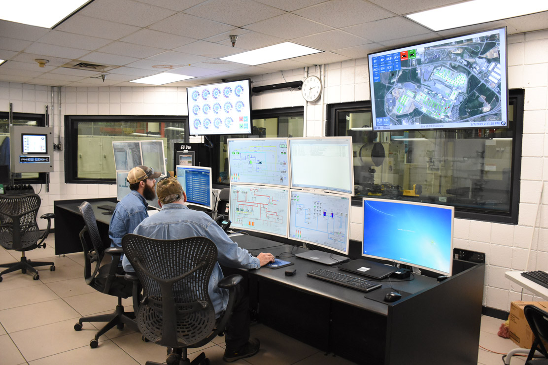 Two men sit at a workstation with multiple monitors displaying technical data and an aerial map.