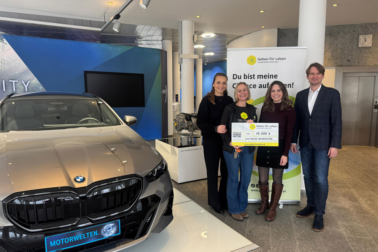 Vier Personen stehen neben einem grauen BMW in einem Ausstellungsraum und halten einen großen symbolischen Scheck mit dem Text 'Geben für Leben'.