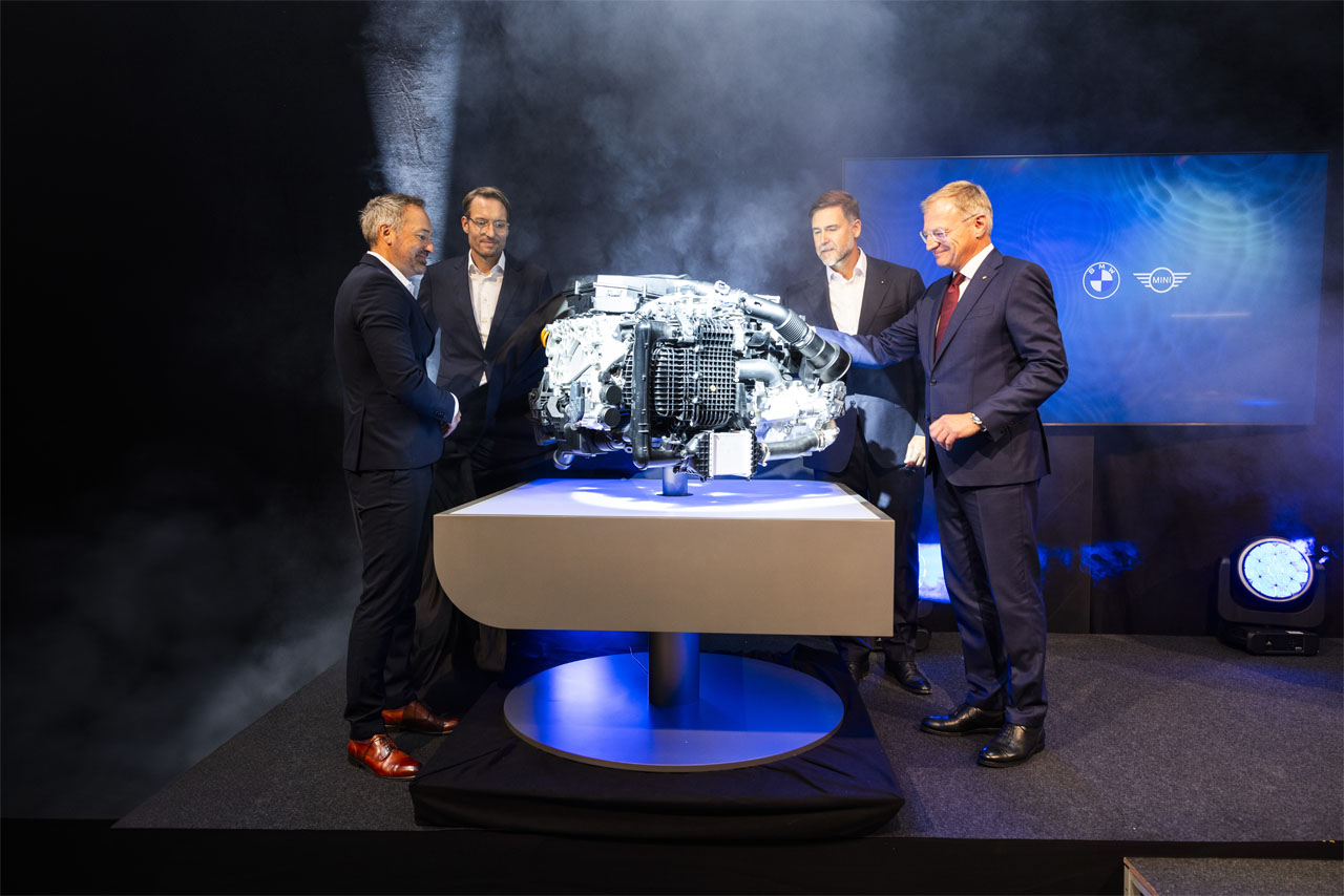 Vier Männer in Anzügen stehen um einen beleuchteten Motor auf einem Podest in einem dunklen Raum mit Nebeleffekt und BMW-Logo im Hintergrund.