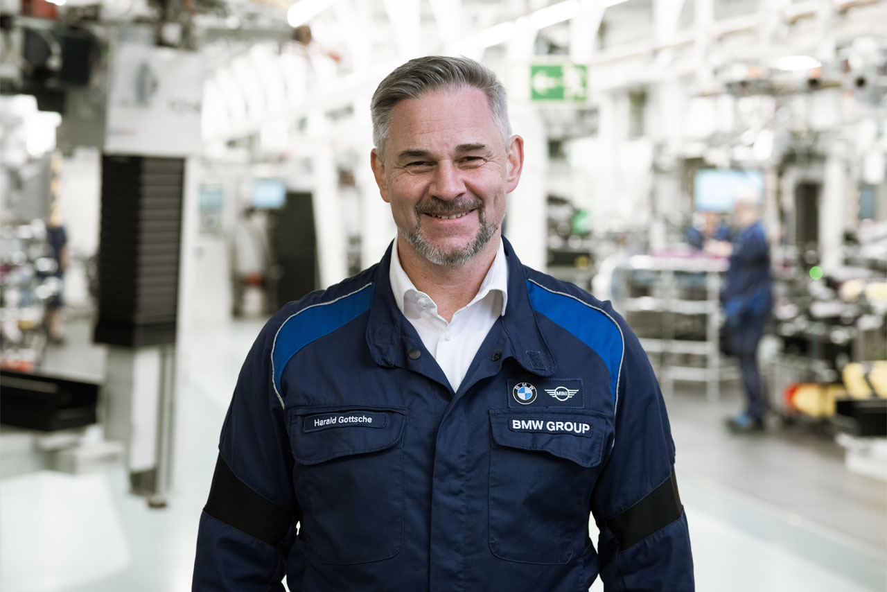 Person in Arbeitskleidung mit Namensschild 'Harald Gottsche' und 'BMW Group' auf der Brust in einer Fabrikumgebung.