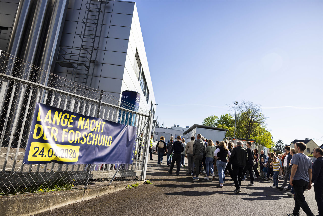 Menschen stehen in einer Schlange vor einem modernen Gebäude mit Zaun und Banner 'LANGE NACHT DER FORSCHUNG 24.04.2026'