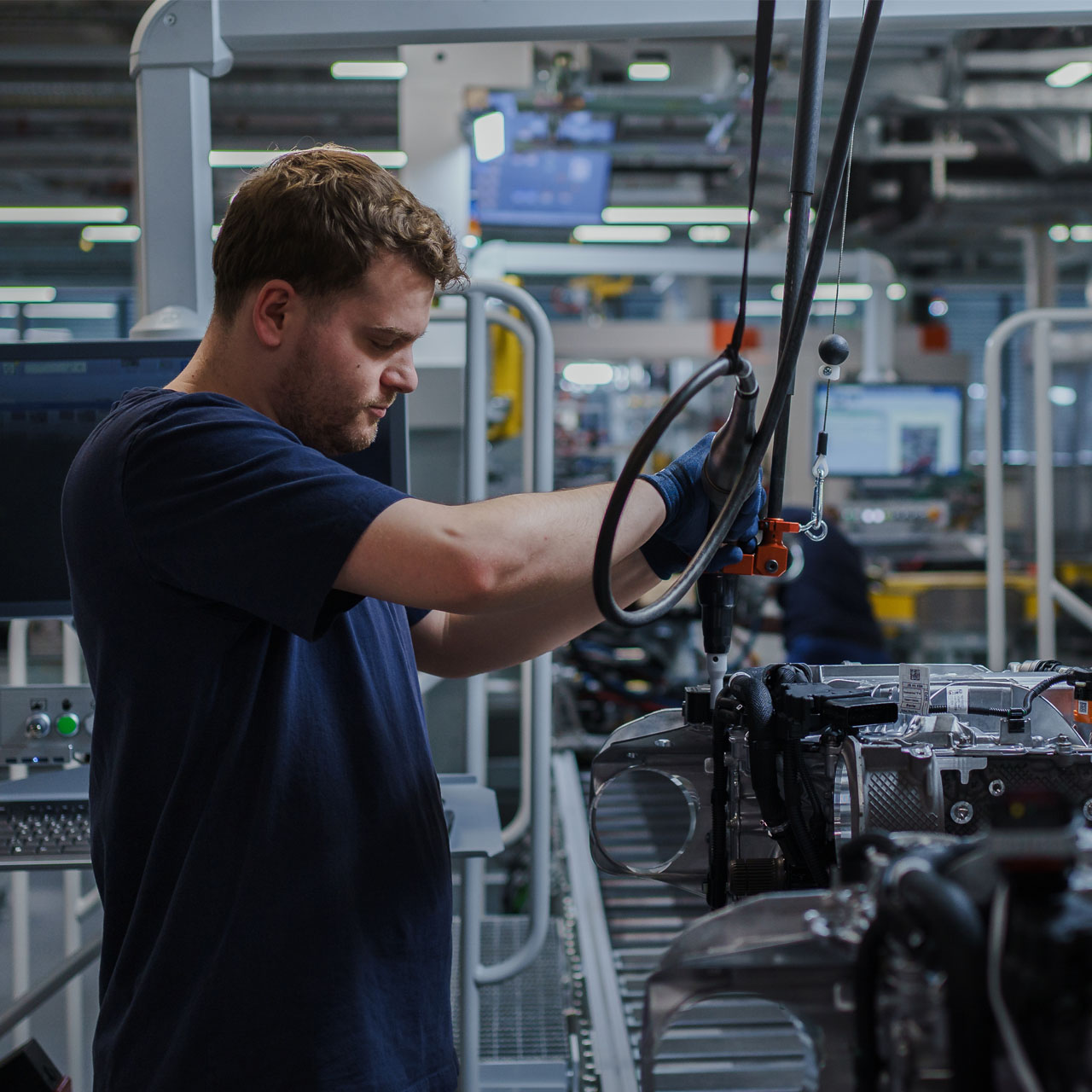 Mann in dunkelblauem T-Shirt arbeitet an einer Maschine in einer Fabrikumgebung mit mehreren technischen Geräten im Hintergrund.