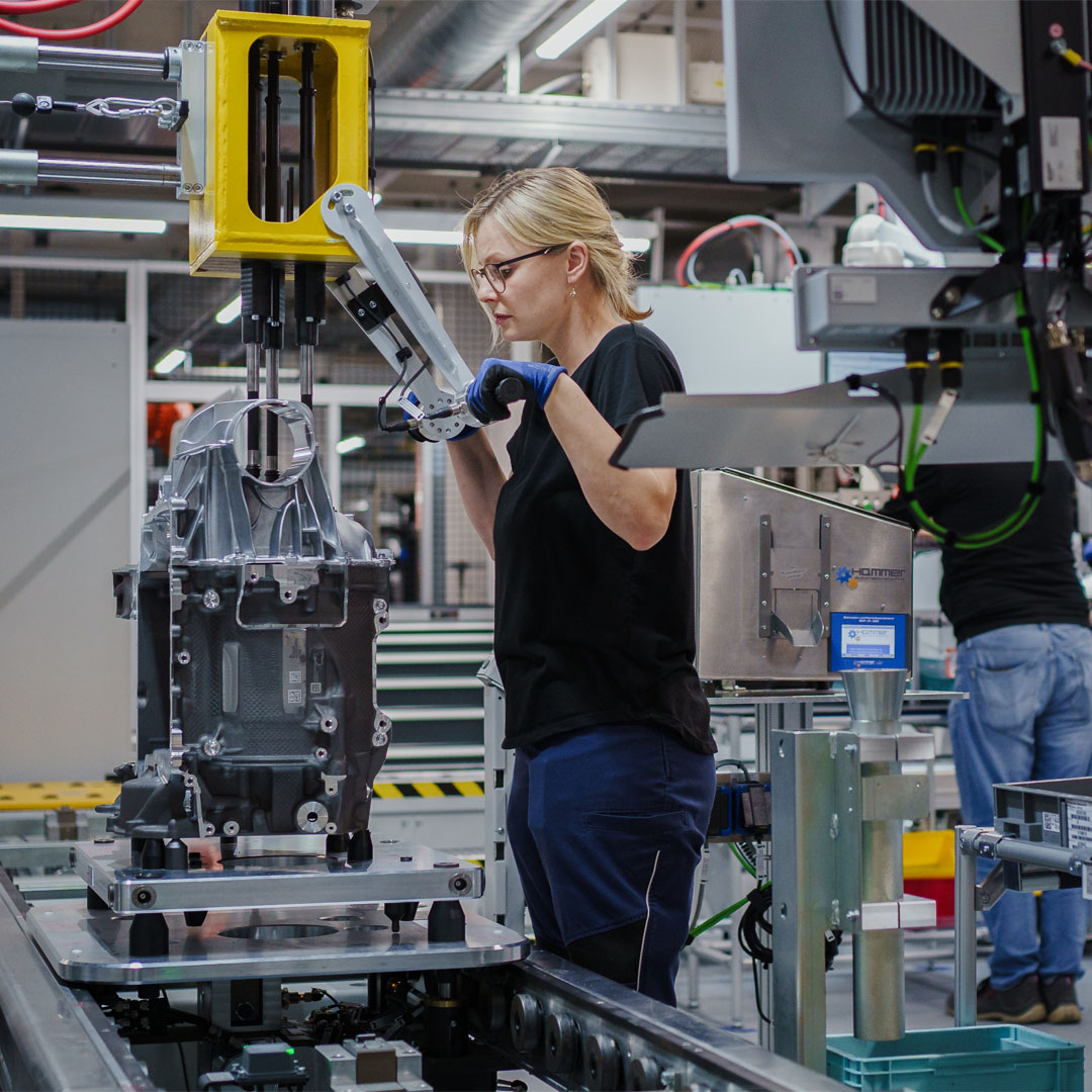 Frau mit Brille und blauen Handschuhen bedient in einer Fabrikhalle eine Maschine mit einem großen Metallteil.