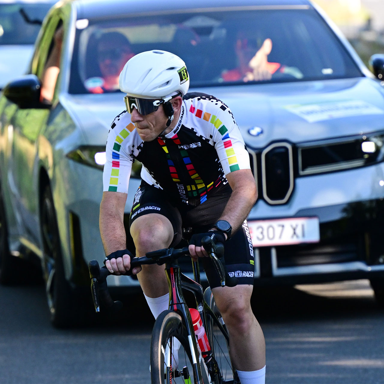 Radfahrer mit weißem Helm und schwarz-weißem Trikot mit bunten Quadraten fährt vor einem schwarzen BMW iX3 auf einer Straße.
