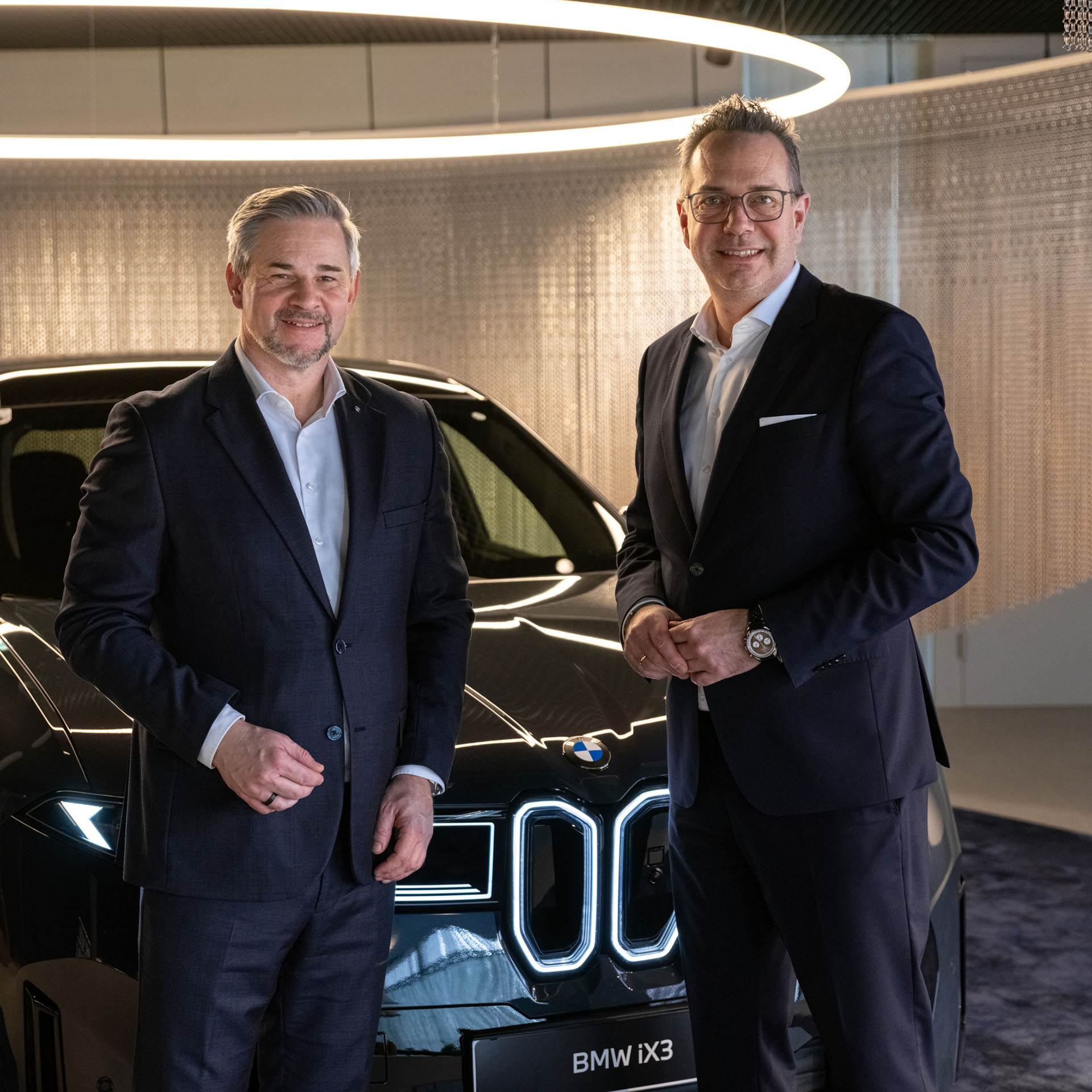 Zwei Männer in Anzügen stehen vor einem schwarzen BMW in einem modernen Innenraum mit rundem Deckenlicht.