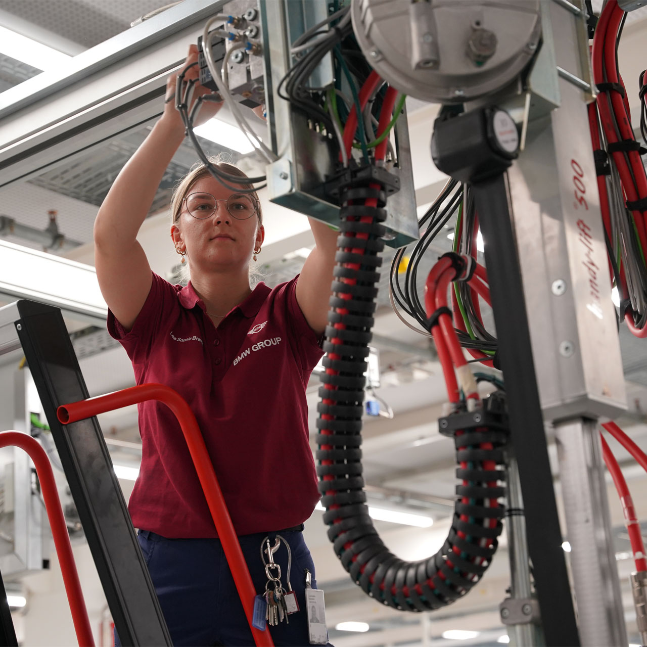 Person in rotem Poloshirt arbeitet an einer technischen Anlage mit Kabeln und Metallteilen in einer Werkhalle.