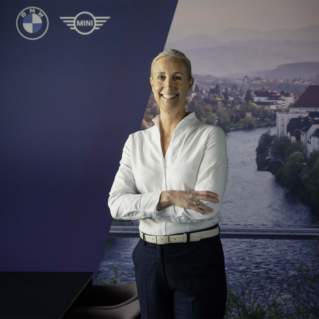 Frau mit weißem Hemd und dunkler Hose steht vor einer Wand mit BMW- und MINI-Logos sowie einem Fluss und einer Stadt im Hintergrund.