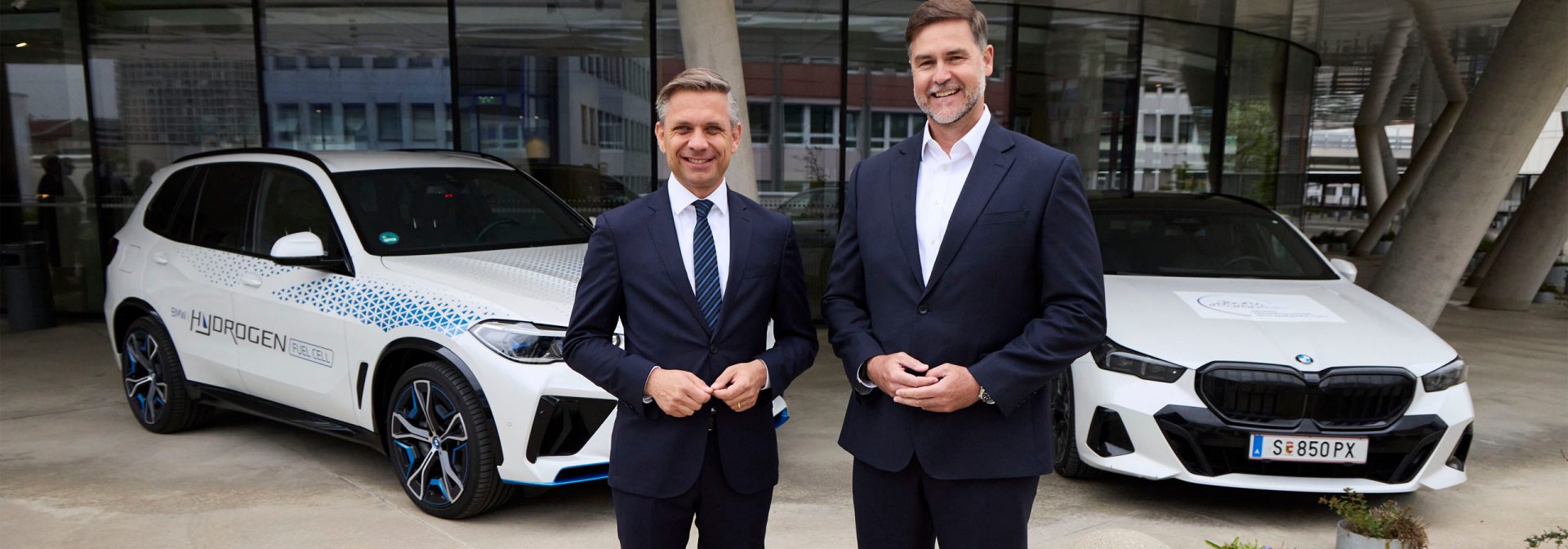 Zwei Männer in eleganten Anzügen stehen neben zwei BMW-Fahrzeuge.