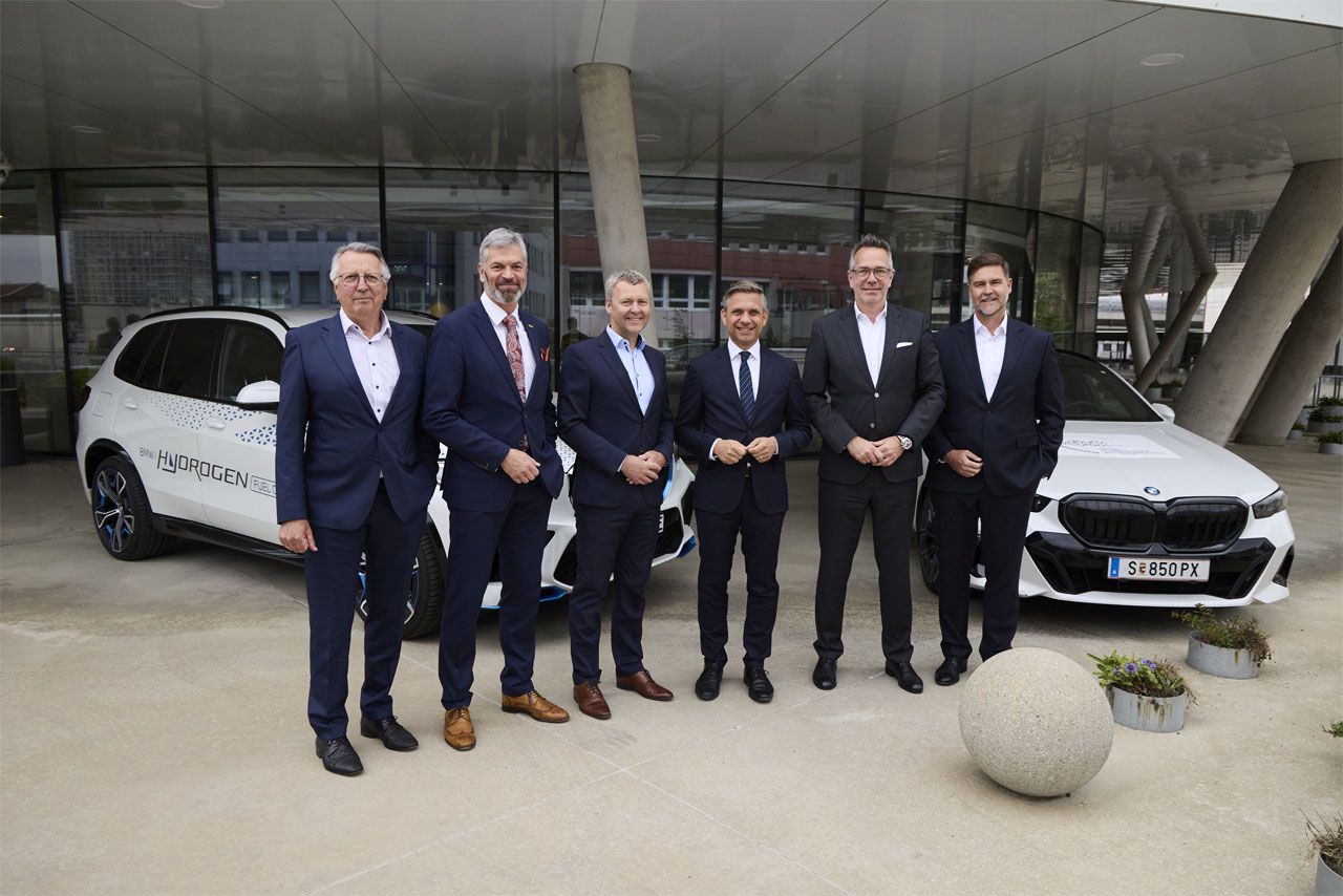 Eine Gruppe von Männern in Anzügen steht vor zwei BMW-Fahrzeugen.