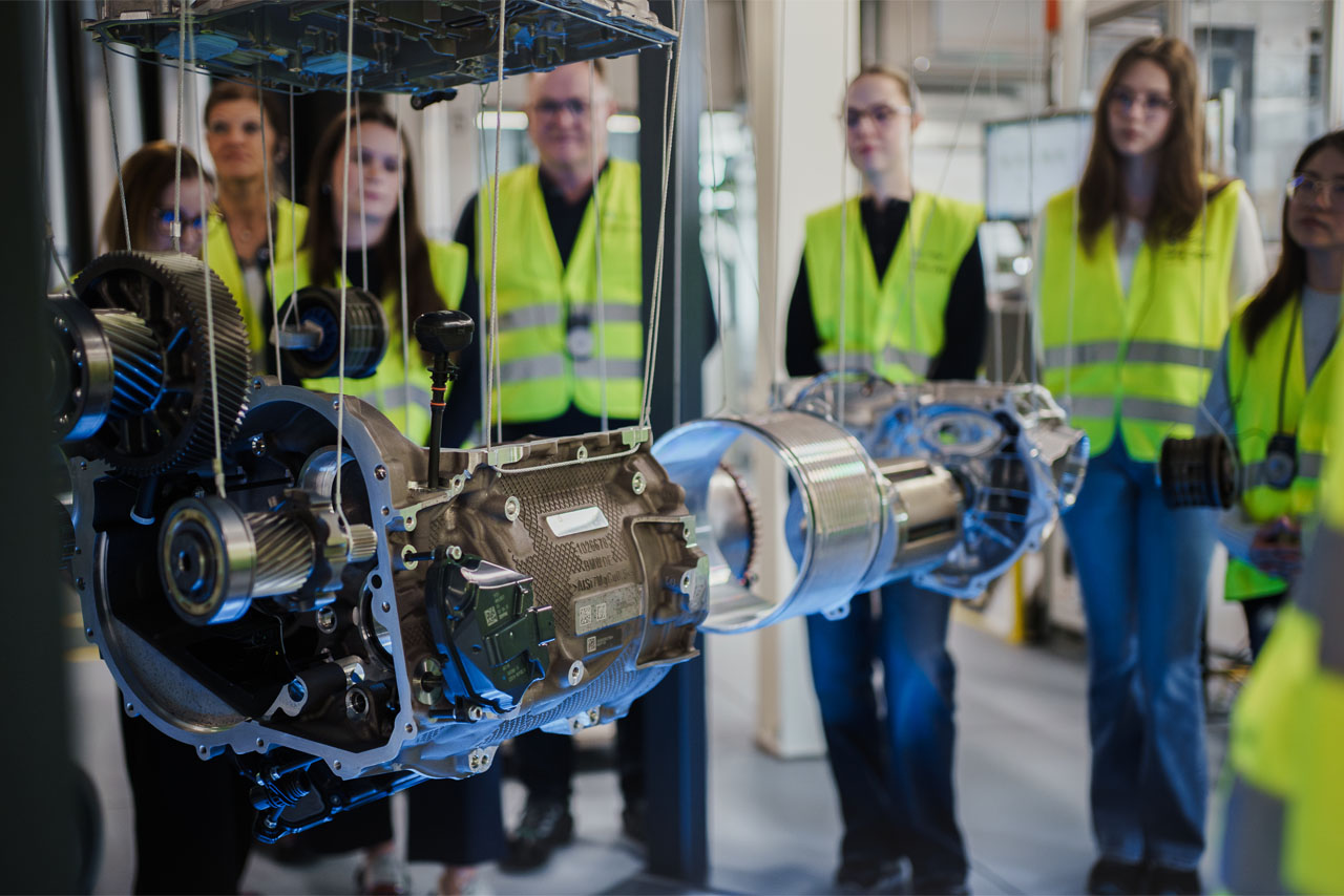 Several people wearing safety vests are standing behind suspended mechanical components in a factory setting.