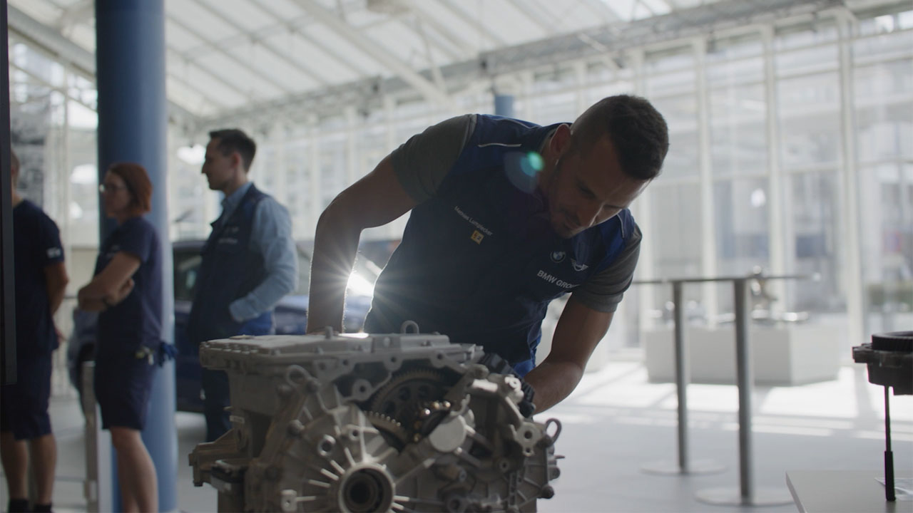 Man leans over an engine in a bright factory hall with other people in the background.