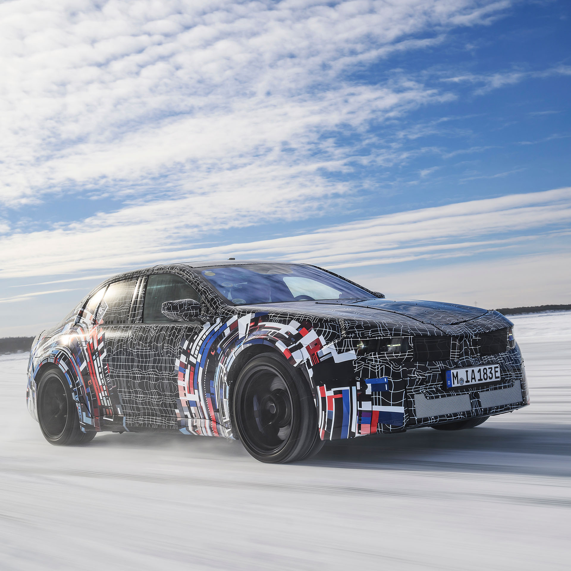 BMW sedan with camouflage wrap in black, white, red, and blue driving on snowy road under cloudy sky.