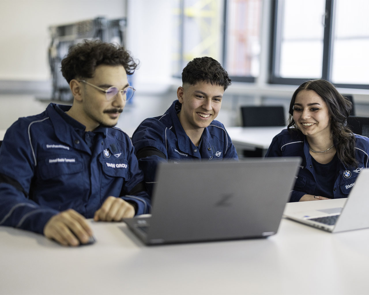 Three people wearing blue work jackets are sitting at a table, looking at two open laptops.