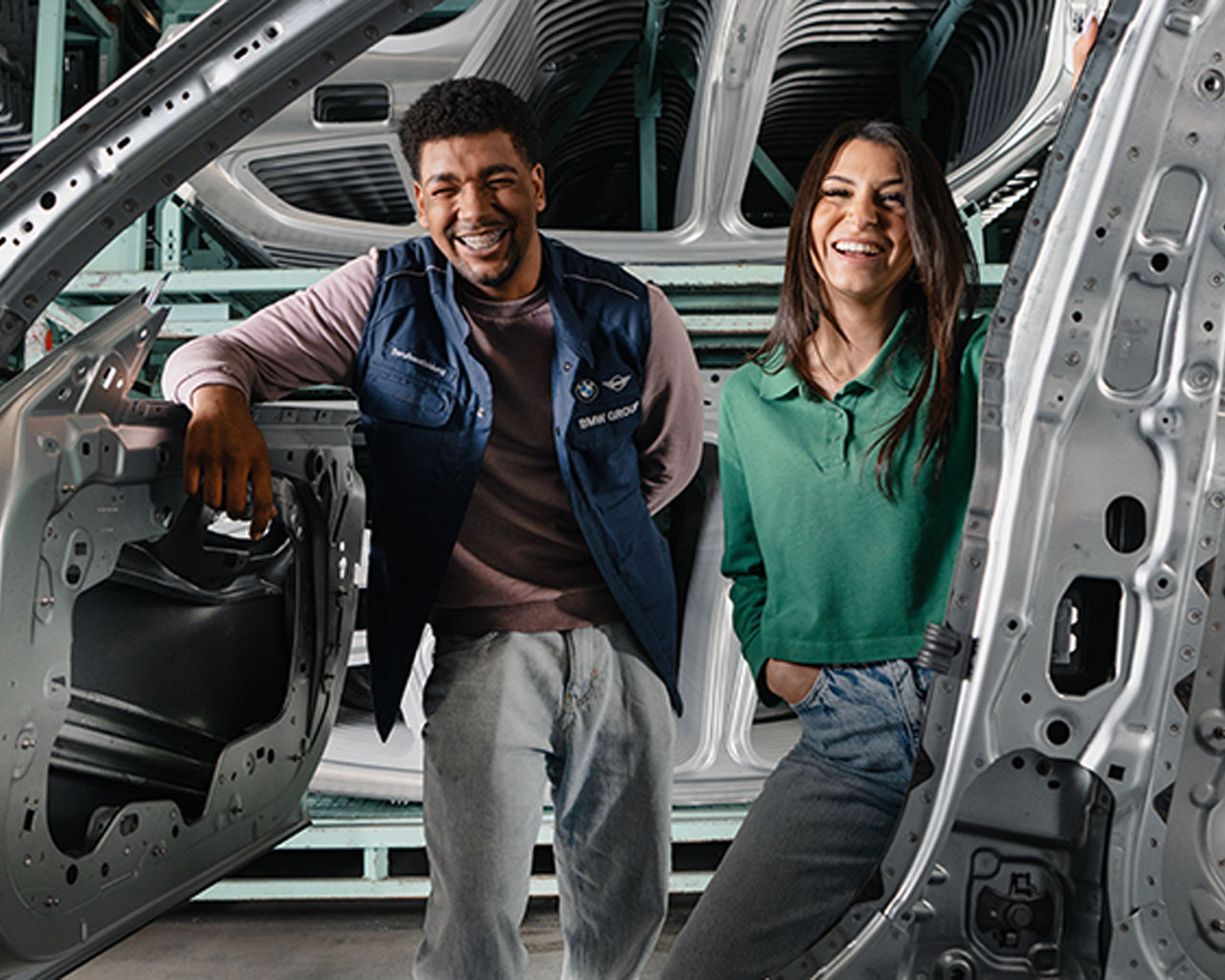 Zwei Auszubildende lächeln durch eine Fahrzeugkarosserie hindurch.